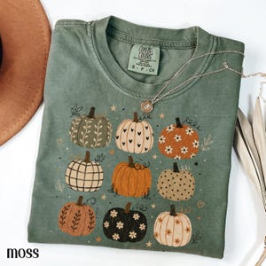 May include: A moss green short-sleeve t-shirt featuring a graphic print of nine pumpkins in various colours and patterns, including floral, heart, and plaid designs. The shirt has a Comfort Colors brand label at the inner neckline and a crew neck.