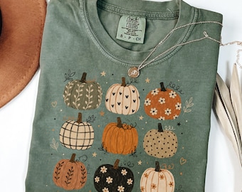 Puede incluir: Una camiseta de manga corta color verde musgo con un estampado gráfico de nueve calabazas en varios colores y patrones, incluyendo corazones, flores y rayas. La camiseta tiene cuello redondo y una etiqueta de la marca Comfort Colors.