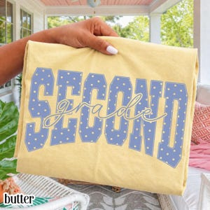 May include: A pale yellow t-shirt featuring the word SECOND in large, blocky, periwinkle blue letters with a white polka dot pattern, overlaid with the word grade in a cursive, light yellow script font across the center of the design.
