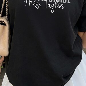 May include: A short-sleeved crew neck t-shirt available in coral, black, or teal. The shirt features the text "FOURTH GRADE" in a bold, sans-serif font, with a personalized name written in a cursive script underneath, such as "Mrs. Johnson" or "Mrs. Taylor."