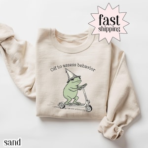 Op de afbeelding: Zandkleurige sweatshirt met een illustratie van een groene kikker met een tovenaars hoed op een scooter. De tekst "Off to assess behavior" staat boven de kikker. De sweatshirt heeft een ronde hals en lange mouwen.