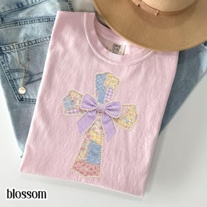 以下が含まれることがあります： パッチワークの十字架のデザインが施された、薄いピンク色のTシャツ。ラベンダーギンガムのリボンとパールの装飾が施されています。「blossom」の文字が下部にプリントされています。ベージュの帽子とブルージーンズが背景にあります。