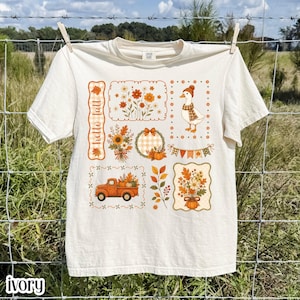 May include: An ivory short-sleeve t-shirt featuring a multi-panel graphic print with the text "hello fall". The design includes a duck wearing a scarf, a vintage truck carrying pumpkins, floral arrangements, autumn leaves, and a festive bunting banner.