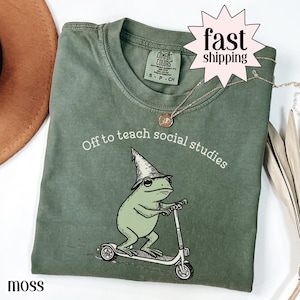 Op de afbeelding: Mosgroen t-shirt met de tekst "Off to teach social studies" en een afbeelding van een kikker op een scooter met een tovenaars hoed. Het shirt heeft een ronde hals en korte mouwen.
