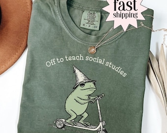 May include: Moss green t-shirt with the text "Off to teach social studies" and a graphic of a frog riding a scooter and wearing a wizard hat. The shirt has a crew neck and short sleeves.