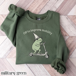 Military green graphic sweatshirt featuring an illustrated frog wearing a wizard hat riding a scooter with “Off to improve mobility” text, whimsical cartoon design