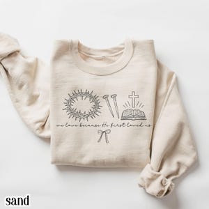 May include: Sand-colored sweatshirt with a crew neck. The front features a black line drawing of a crown of thorns, nails, a cross, an open book, and a bow. The text "we love because He first loved us" is below the drawings. The word "sand" is at the bottom.