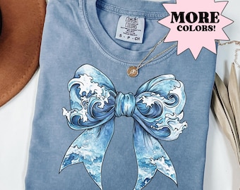 May include: A blue t-shirt with a graphic of a blue bow made of ocean waves. The shirt has the text "Comfort Colors" and "S - P - CH" printed on the tag.