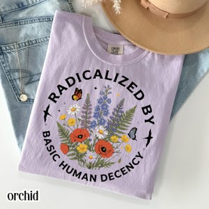 Pode incluir: Camiseta cor de orquídea com um design floral e o texto "RADICALIZED BY BASIC HUMAN DECENCY" em disposição circular. O design inclui borboletas, flores e estrelas. A camiseta está dobrada sobre jeans.