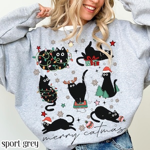Pode incluir: Camisola cinza desportiva com um design natalício. Apresenta gatos pretos adornados com elementos festivos como luzes, enfeites e gorros de Pai Natal. As palavras "merry catmas" estão escritas em itálico.