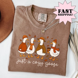 Espresso brown Comfort Colors t-shirt featuring illustrated geese in cozy fall outfits with scarves and hats, autumn leaves, and the phrase “just a cozy goose,” cute goose graphic shirt, fall aesthetic shirt, cozy autumn tee, whimsical animal design, soft unisex shirt, fall gift idea.