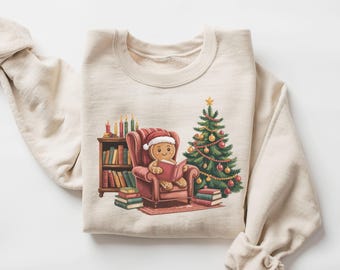 May include: Sand-coloured sweatshirt with a gingerbread man in a Santa hat reading a book in a red armchair. A Christmas tree and bookshelf are to the right. The word "sand" is printed at the bottom.