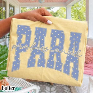 Könnte beinhalten: Ein blassgelbes T-Shirt mit dem Wort PARA in großen, blockartigen, pervenche-blauen Buchstaben mit gelbem Punktmuster. Das Wort professional ist in einer kursiven, pervenche-blauen Schreibschrift über die Blockbuchstaben gelegt.