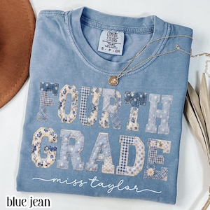 May include: A blue jean Comfort Colors t-shirt with the words "FOURTH GRADE" in patchwork letters. The letters are decorated with floral and gingham patterns. The name "miss taylor" is written in cursive below the words. A gold necklace rests on the shirt.