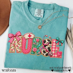 Pode incluir: Uma t-shirt de manga curta azul-petróleo com uma estampa gráfica que soletra a palavra NURSE. As letras apresentam padrões como xadrez rosa, flores, bolinhas e manchas de vaca. Inclui um estetoscópio rosa, uma touca de enfermeira e dois laços.