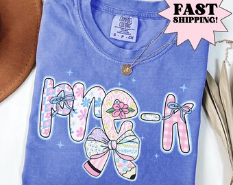 Peut inclure: Un t-shirt bleu pervenche avec le texte "Pre-K" dans une police fantaisiste, décoré de motifs floraux et de nœuds. Un collier en or avec un pendentif est drapé sur le t-shirt. Le t-shirt porte une étiquette "Comfort Colors".