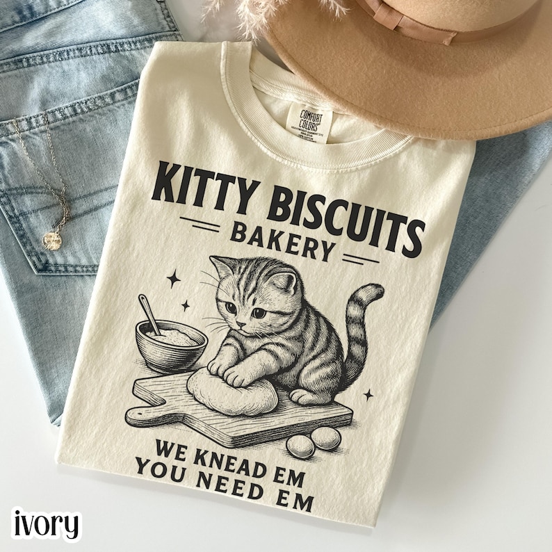 Ivory graphic t-shirt featuring a baking cat illustration kneading dough with “Kitty Biscuits Bakery” and “We knead em you need em” text, cat bakery shirt