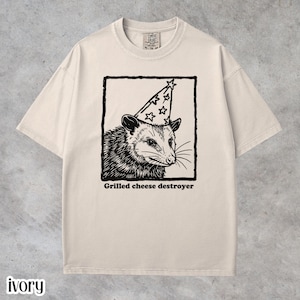 Ivory Comfort Colors unisex t-shirt featuring a quirky black line-art illustration of a possum wearing a party hat inside a framed graphic with the text “Grilled cheese destroyer.” Funny possum graphic tee, vintage-style humor shirt, soft-washed cotton unisex t-shirt, foodie joke apparel, animal meme shirt, casual everyday wear, and unique gift for possum lovers and humor graphic tee fans