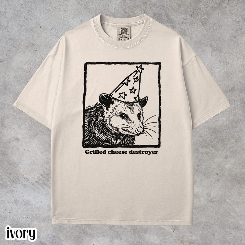 Ivory Comfort Colors unisex t-shirt featuring a quirky black line-art illustration of a possum wearing a party hat inside a framed graphic with the text “Grilled cheese destroyer.” Funny possum graphic tee, vintage-style humor shirt, soft-washed cotton unisex t-shirt, foodie joke apparel, animal meme shirt, casual everyday wear, and unique gift for possum lovers and humor graphic tee fans