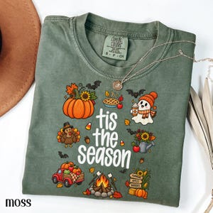 Könnte beinhalten: Ein moosgrünes Kurzarm-T-Shirt mit dem weißen Schriftzug 'tis the season', umgeben von bunten herbstlichen Illustrationen wie einem Kürbis, einem Truthahn, einem Geist, einem roten Lastwagen, einem Lagerfeuer und einem Schild 'pumpkins apples hayrides'.