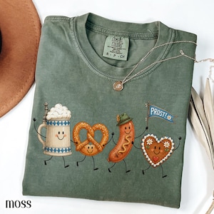 May include: A moss green short-sleeve t-shirt featuring a graphic of a beer stein, a pretzel, a sausage wearing a hat, and a heart-shaped cookie holding a blue flag with the text PROST!. The shirt has a Comfort Colors brand neck label.