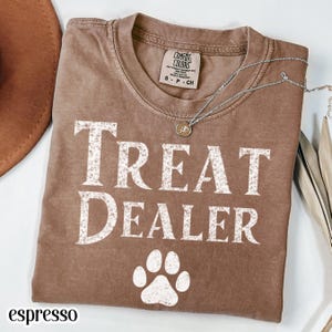 Espresso brown Comfort Colors t-shirt featuring distressed white typography reading treat dealer with a paw print graphic below. Soft-washed cotton short sleeve graphic tee with playful pet lover humor, vintage style lettering, rustic neutral tone, and casual dog mom aesthetic