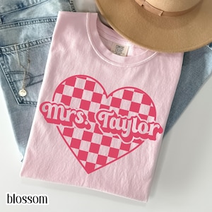 May include: A light pink t-shirt with a large heart-shaped design. The heart features a pink and white checkered pattern and the text "Mrs. Taylor" in a retro font. The word "blossom" is printed at the bottom.