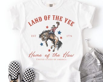 May include: White t-shirt with a graphic of a cowboy riding a bucking bronco, with text that reads "Land of the Yee" and "Home of the Haw". The design includes red and blue stars. Black and white checkered slip-on shoes are at the bottom.