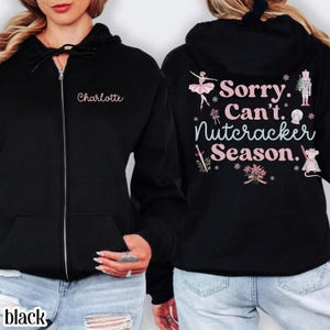 Könnte beinhalten: Ein schwarzer Kapuzenpullover mit Reißverschluss und dem Namen Charlotte in Pink auf der linken Brust. Auf der Rückseite befindet sich ein Aufdruck mit dem Text Sorry. Can't. Nutcracker Season. sowie Illustrationen einer Ballerina und Nussknacker.