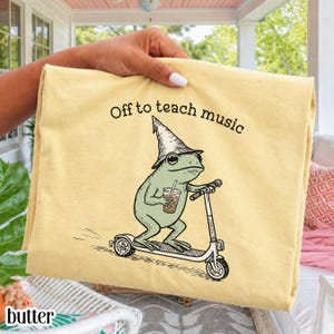 May include: A pale yellow t-shirt featuring a graphic of a green frog wearing a pointed hat, riding a scooter, and holding a coffee cup. The text above the frog reads "Off to teach music" in a simple black font. The design is centered on the fabric.