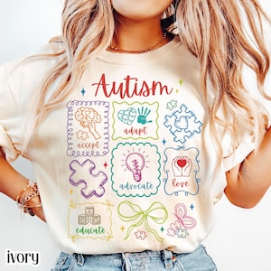 Puede incluir: Camiseta de color marfil con la palabra "Autismo" en escritura roja. La camiseta presenta cuadrados gráficos coloridos con palabras como "aceptar", "adaptar" y "amar", junto con piezas de rompecabezas y una bombilla.