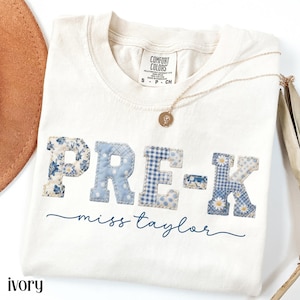 May include: Ivory-colored t-shirt with the text "PRE-K miss taylor" in blue patchwork letters. The letters are decorated with floral, gingham, and daisy patterns. A gold necklace with a pendant is draped on the shirt.