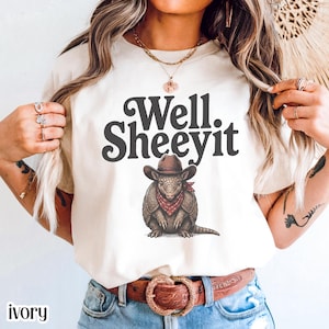 May include: Ivory t-shirt with the text "Well. Sheeeyit" and an illustration of an armadillo in a cowboy hat and red bandana. The armadillo is shaded in brown and tan. The shirt has a crew neck and is made of a soft material.