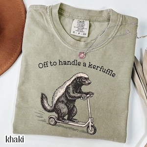 Khaki graphic t-shirt featuring a vintage illustration of a honey badger riding a scooter with the phrase “Off to handle a kerfuffle,” funny honey badger shirt, quirky animal humor tee, whimsical vintage-style artwork, unisex comfort color t-shirt, novelty graphic apparel, unique gift for animal lovers and humor fans