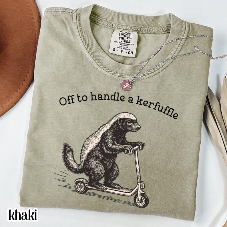 Khaki graphic t-shirt featuring a vintage illustration of a honey badger riding a scooter with the phrase “Off to handle a kerfuffle,” funny honey badger shirt, quirky animal humor tee, whimsical vintage-style artwork, unisex comfort color t-shirt, novelty graphic apparel, unique gift for animal lovers and humor fans