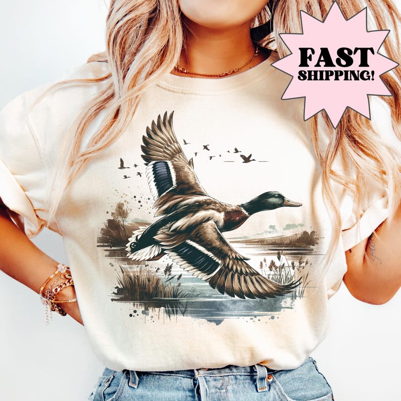Ivory graphic t-shirt featuring a flying duck illustration over a marsh landscape with water and birds in the background, detailed wildlife artwork