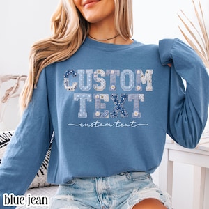 Puede incluir: Una sudadera azul de manga larga con cuello redondo que presenta las palabras CUSTOM TEXT en una fuente tipo patchwork con varios patrones florales y de cuadros vichy en azul y blanco. Debajo, las palabras custom text en letra cursiva blanca.