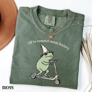 Può includere: Una maglietta verde muschio con la scritta "Off to commit mom foolery" e un disegno di un ranocchio con un cappello da mago su un monopattino. Una collana d'oro è drappeggiata sulla maglietta.