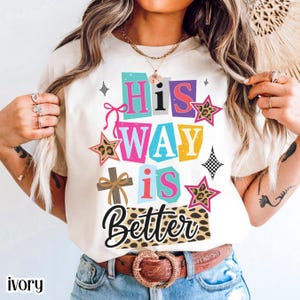 May include: Ivory-colored t-shirt with the text "His WAY is Better" in colorful, block letters. The design includes a cross, stars with leopard print, and decorative elements. The shirt has a crew neck and short sleeves.