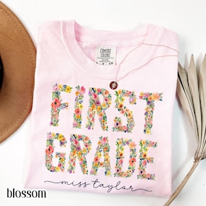 May include: A light pink t-shirt with the words "FIRST GRADE" in colorful floral letters. The shirt also features the text "miss taylor" in cursive and the word "blossom" in a bold font. A gold necklace with a pendant is draped on the shirt.