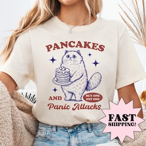 May include: A white t-shirt featuring a blue and white illustration of a cat holding a stack of pancakes. The text "Pancakes and Panic Attacks" is printed on the shirt with a red "Buy One Get One" tag.