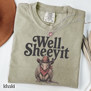 May include: Khaki-coloured t-shirt featuring the text "Well Sheeeyit" in dark brown. An illustration of an armadillo wearing a cowboy hat and bandana is below the text. A silver necklace with a pendant is on the shirt.