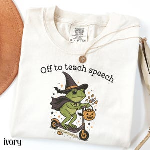 Può includere: T-shirt color avorio con la scritta "Off to teach speech". La maglietta presenta una rana verde con cappello e mantello da strega, che guida un monopattino e porta un secchio di zucca. Una collana d'oro è drappeggiata sulla maglietta.