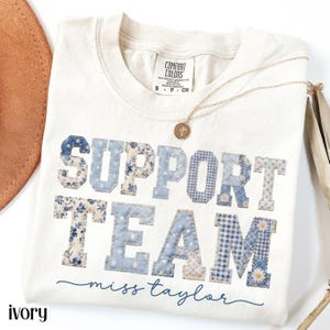 May include: Ivory-colored t-shirt with the words "SUPPORT TEAM" in blue patchwork letters. The shirt also features the name "miss taylor" in cursive and a gold necklace. The shirt is a Comfort Colors brand.