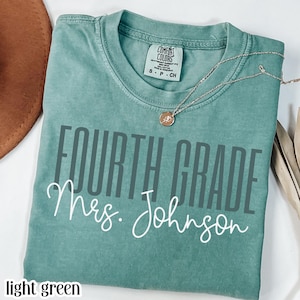 May include: A light green short-sleeve t-shirt featuring the text "FOURTH GRADE" in a dark gray sans-serif font and "Mrs. Johnson" in a white cursive script. The shirt has a Comfort Colors brand label at the neck and a thin gold-tone necklace.