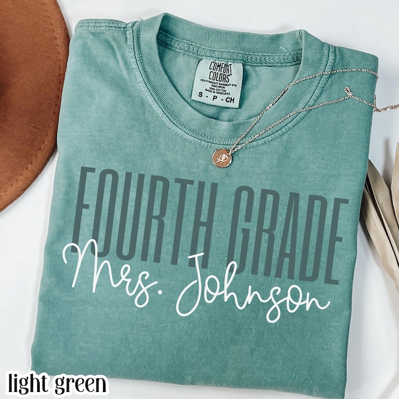 May include: A light green short-sleeve t-shirt featuring the text "FOURTH GRADE" in a dark gray sans-serif font and "Mrs. Johnson" in a white cursive script. The shirt has a Comfort Colors brand label at the neck and a thin gold-tone necklace.