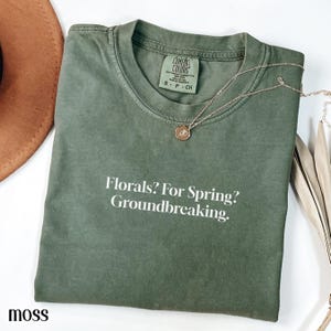 May include: A moss green t-shirt with the text "Florals? For Spring? Groundbreaking." A gold necklace with a pendant rests on the shirt. The shirt is folded and has a tag that says "Comfort Colors".