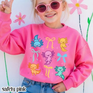 May include: A safety pink sweatshirt with a crew neck, decorated with cartoon axolotls and bows in various colours. The axolotls are blue, yellow, pink, and green. The text "safety pink" is at the bottom. A bright and cheerful design.