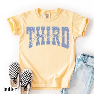 May include: A pale yellow short-sleeve t-shirt featuring the word THIRD in large blue block letters with white polka dots, overlaid with the word grade in a white cursive script. The shirt has a crew neckline and a Comfort Colors brand label.