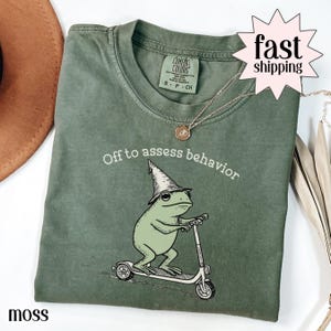 Op de afbeelding: Mosgroen t-shirt met een afbeelding van een kikker met een tovenaars hoed en op een scooter. De tekst "Off to assess behavior" staat boven de kikker. Een gouden ketting is zichtbaar.